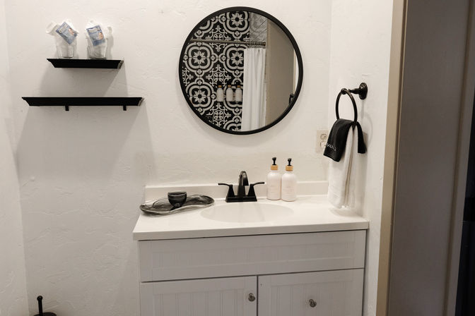 Small white bathroom vanity with black faucet, round black-framed mirror reflecting patterned shower tile, wall towel ring, floating shelves and soap dispensers