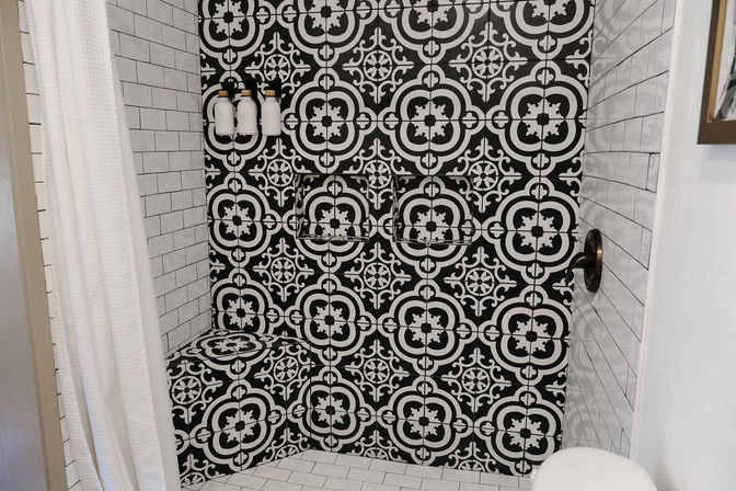 Black-and-white patterned tile shower with built-in bench, white subway tile surround, wall-mounted soap dispensers and bronze shower handle.