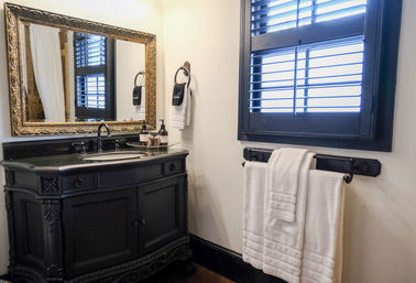 Elegant powder room with ornate black vanity, gold-framed mirror, black shuttered window and neatly folded white towels on a wall rack