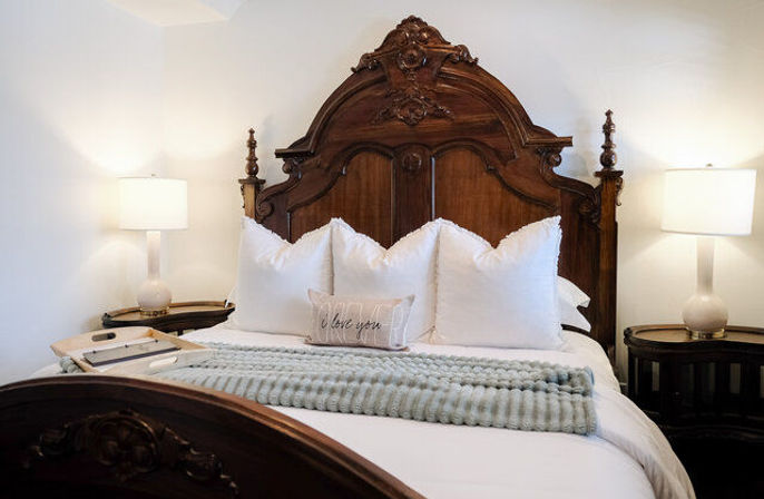 Cozy elegant bedroom interior with ornate carved wooden headboard, crisp white pillows, 'I love you' accent pillow, soft knit throw and matching bedside lamps.