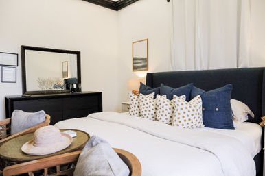 Cozy modern bedroom interior with navy upholstered headboard, crisp white bedding, row of blue and gold polka-dot throw pillows, black dresser with large mirror, bedside lamp, and wicker chairs with a straw sun hat.