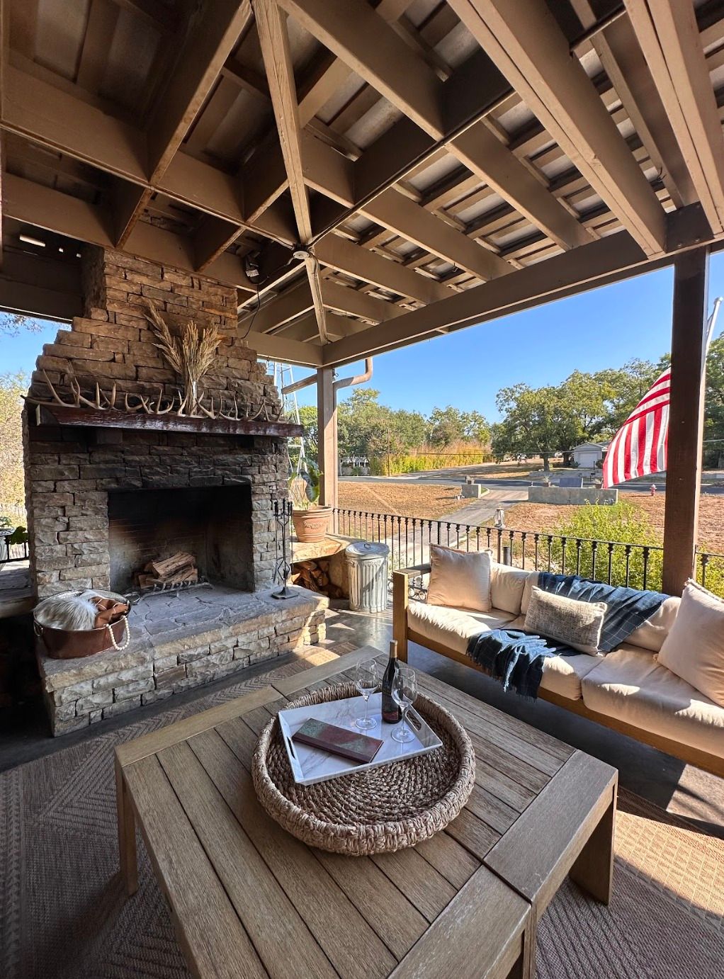 Cozy covered outdoor patio with stone fireplace, wooden coffee table topped with wine and glasses, cushioned sofa and throw blanket, and an American flag overlooking a rural yard and trees under a blue sky.