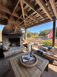 Cozy covered outdoor patio with stone fireplace, wooden coffee table topped with wine and glasses, cushioned sofa and throw blanket, and an American flag overlooking a rural yard and trees under a blue sky.