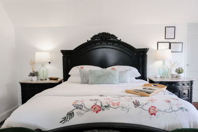 Cozy master bedroom with ornate black headboard, white floral-embroidered duvet, pastel throw pillows, matching nightstands with glass lamps and a wooden tray on the bed.