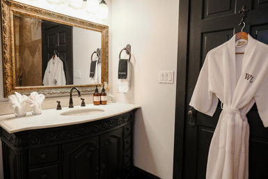 Cozy luxury bathroom interior with ornate gold mirror, marble countertop over carved dark wood vanity, amber soap bottles and a white waffle robe hanging on a black door