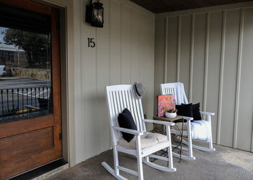 Inviting front porch with two white wooden rocking chairs with cushions and throws, a small metal side table holding a potted plant and colorful book, neutral vertical siding, lantern light and wooden front door.