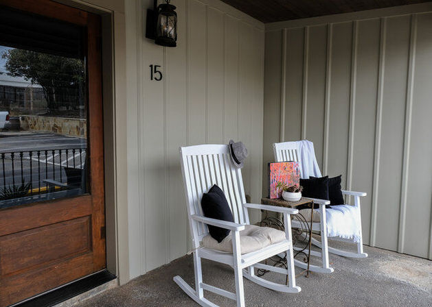 Inviting front porch with two white wooden rocking chairs with cushions and throws, a small metal side table holding a potted plant and colorful book, neutral vertical siding, lantern light and wooden front door.