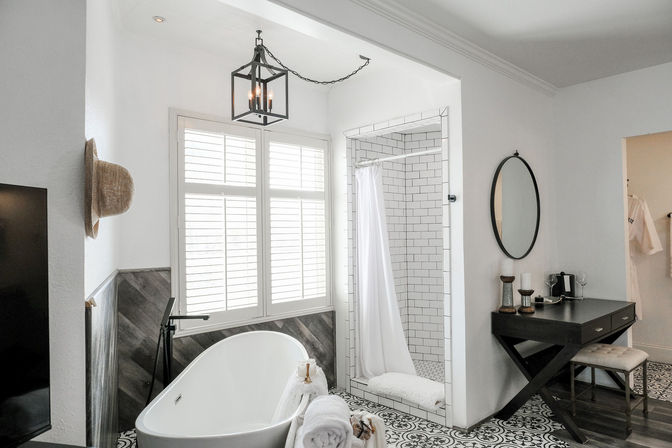 Bright modern farmhouse bathroom with a freestanding oval tub, white subway-tile shower with curtain, black-and-white patterned floor tile, black metal lantern pendant, shuttered window, and dark wood vanity with round mirror.