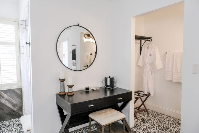 Chic bright bathroom dressing area with a black vanity desk, round wall mirror, cushioned stool, decorative candles and a coffee setup; white robe hanging in an open closet and black-and-white patterned tile floor.