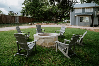 Cozy stone fire pit surrounded by six gray Adirondack chairs on a green backyard lawn, with a gray farmhouse, large oak tree, and wooden fence in the background.