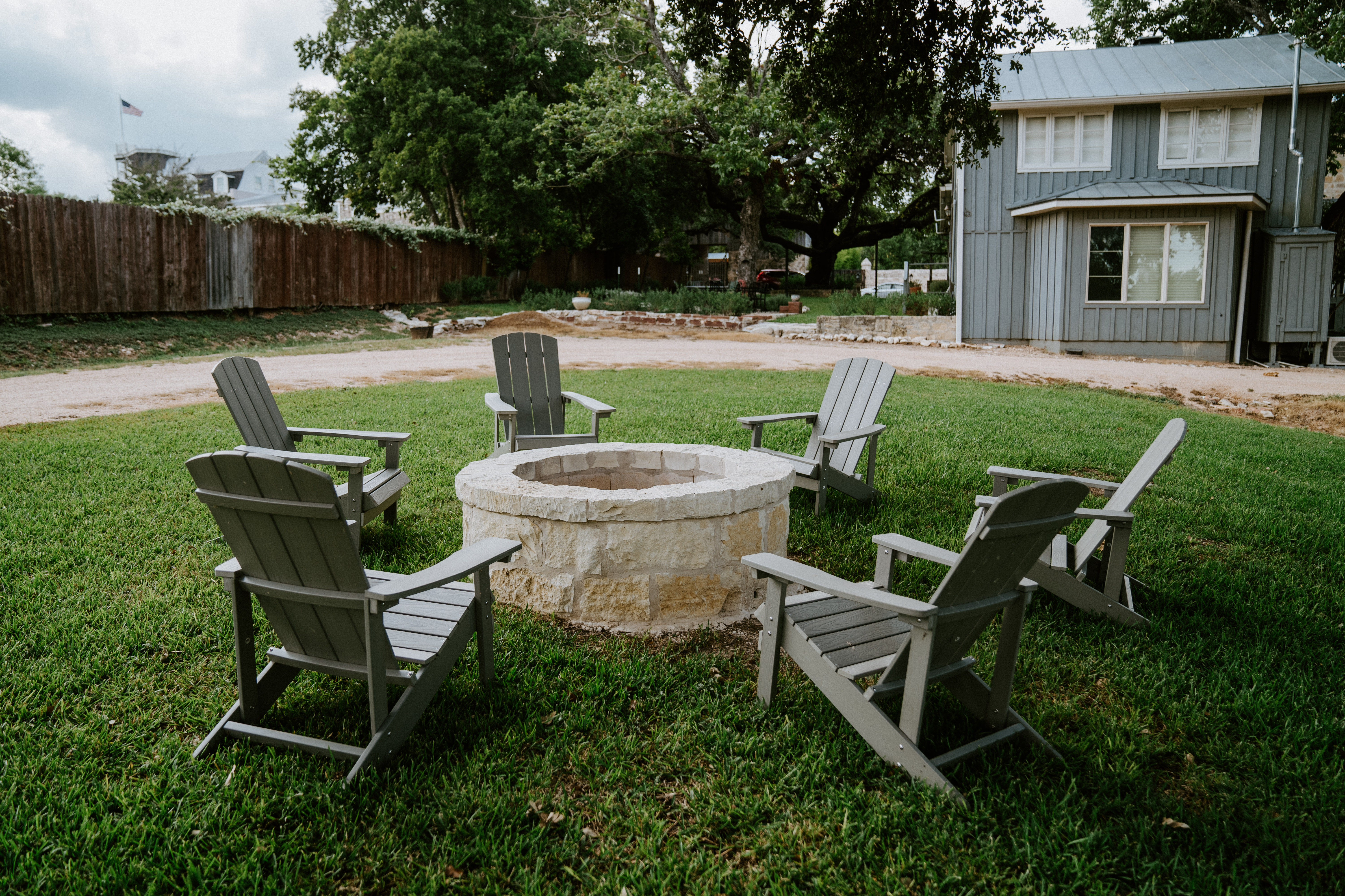 Cozy stone fire pit surrounded by six gray Adirondack chairs on a green backyard lawn, with a gray farmhouse, large oak tree, and wooden fence in the background.