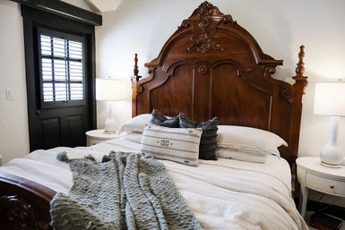 Cozy bedroom with an ornate antique wooden headboard, crisp white bedding, textured gray throw and decorative pillows, round nightstands with lamps, and a black shuttered door.