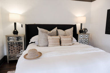 Stylish cozy bedroom with black upholstered headboard, crisp white bedding, layered beige textured pillows, straw sun hat on the bed, mirrored nightstands with black lamps and decorative accents, white walls and dark hardwood floor.