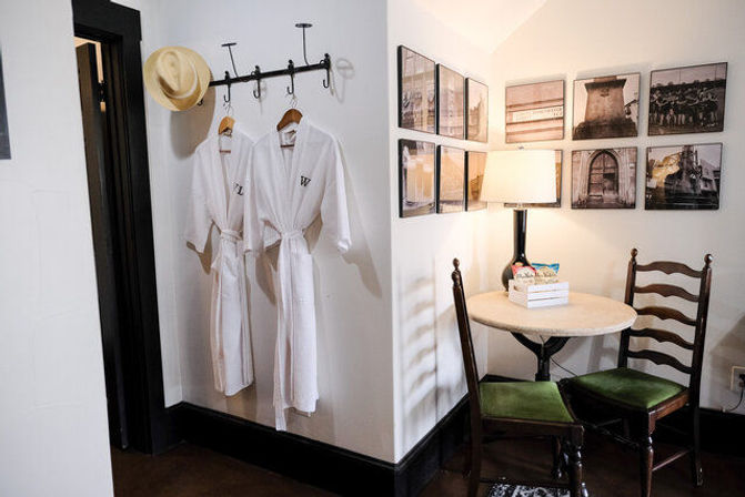 Cozy hotel-room nook with two white monogrammed bathrobes and a straw hat on wall hooks, round marble-top table with lamp and two wooden chairs, and a gallery wall of framed photos.