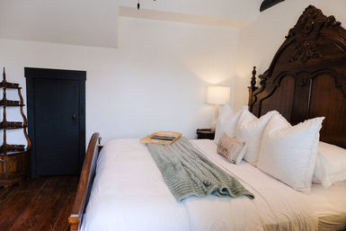 Cozy vintage bedroom with ornate dark-wood headboard, crisp white linens and pillows, sage-green throw and bed tray, bedside lamp and hardwood floor.