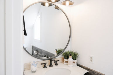 Bright modern bathroom vanity with round wall mirror, oil-rubbed bronze faucet and white countertop, small potted greenery and soap dispenser under metal sconce lights.