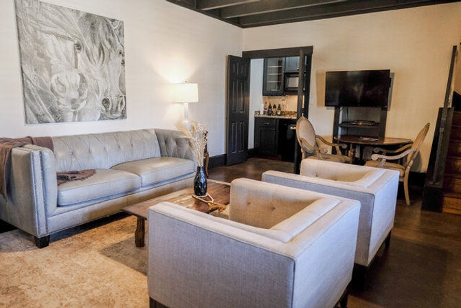 Cozy contemporary living room with a gray tufted sofa, two matching armchairs, wooden coffee table, abstract wall art, floor lamp, flat-screen TV, hardwood floors and a kitchenette visible through an open doorway.