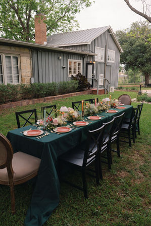 Outdoor farmhouse garden dinner setup — long table on lawn with emerald tablecloth, coral plates, floral centerpieces and candles, black cross-back chairs.