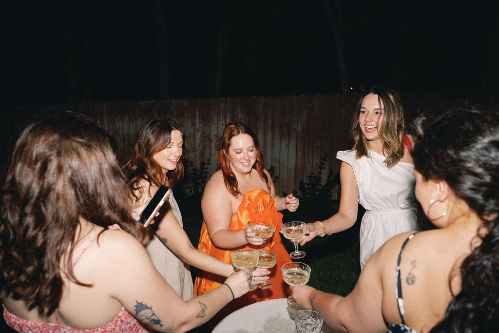Group of friends at a backyard evening party clinking champagne coupe glasses around a small table, smiling and laughing under a dark sky.
