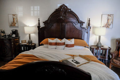 Cozy boutique bedroom with ornate dark-wood headboard, white and rust-orange bedding, three decorative pillows, bedside lamps and vintage side tables
