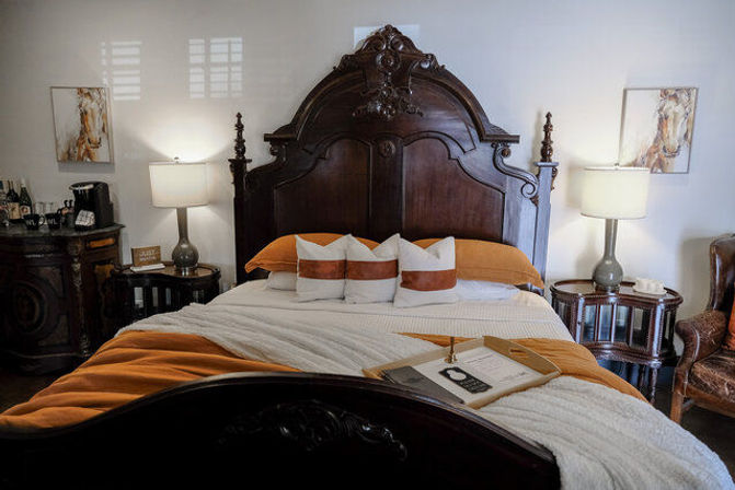 Cozy boutique bedroom with ornate dark-wood headboard, white and rust-orange bedding, three decorative pillows, bedside lamps and vintage side tables