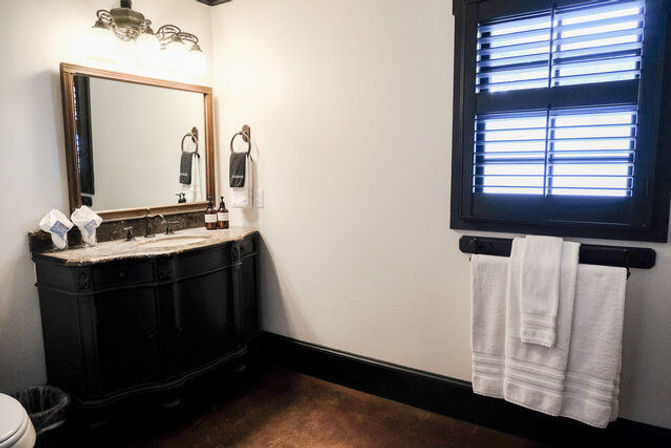 Cozy modern bathroom with dark wood vanity and marble countertop, large framed mirror and bronze light fixture, black plantation shutters, and neatly folded white towels on a wall rack.