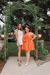 Two smiling women pose under a vine-covered arch with purple flowers in a backyard garden — one in a white ruffled mini dress and black heels, the other in a bright orange strapless dress with a large rosette and white pumps, lanterns flanking the arch and a large tree behind them.