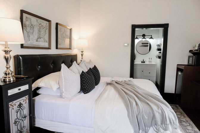 Cozy boutique-style bedroom with tufted dark headboard, layered white bedding, striped and patterned pillows, gray throw, brass bedside lamps, framed maps, and an ensuite bathroom with round mirror.