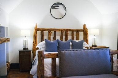 Cozy rustic bedroom with a natural log-frame bed, three navy throw pillows, gray upholstered bench, wooden nightstands with glass table lamps, round wall mirror, white walls and hardwood floor.