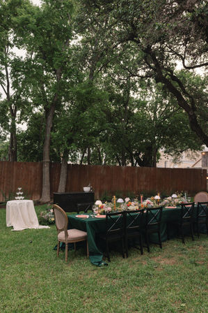 Charming backyard dinner under tall trees — long table on the lawn with a dark green tablecloth, floral centerpieces, small lamps and candles, mixed dining chairs and a white-clothed champagne tower by a wooden fence.