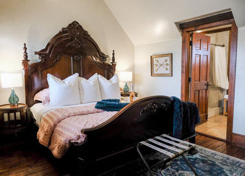 Cozy cottage-style bedroom with ornate carved antique wooden bed, white pillows and pink quilt, teal throw, bedside lamps, hardwood floors and open door to en‑suite bathroom.