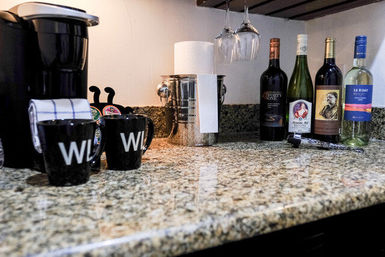 Granite countertop beverage station with single-serve coffee maker, two black mugs, coffee pods and towel, stainless ice bucket, hanging wine glasses and four assorted wine bottles ready for coffee or evening wine.