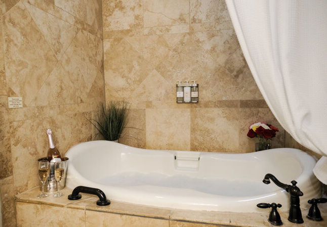 Spa-like white soaking bathtub in beige marble-tiled bathroom with champagne on ice, two glasses, and a vase of roses