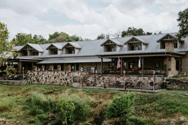 Charming riverside country lodge with metal roof and dormer windows, stone terrace and outdoor patio with potted plants, American and state flags, overlooking a grassy riverbank.