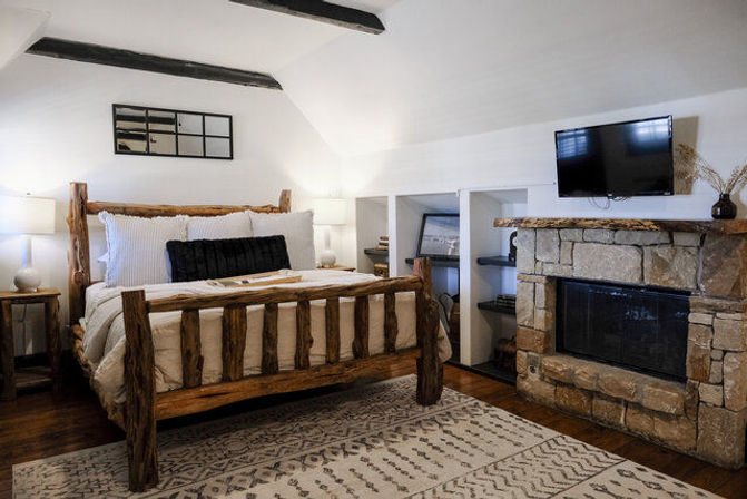 Cozy rustic attic bedroom with log-frame bed, white linens and black accent pillow, stone fireplace with wall-mounted TV, built-in shelves, patterned rug and exposed ceiling beams.