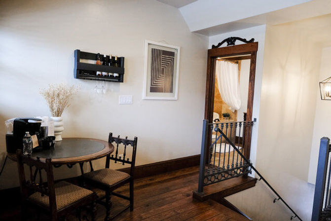 Cozy upstairs landing with round wood table, two carved chairs, a coffee station and dried-flower vase, wall wine rack and framed art, hardwood floors and an ornate doorway to a curtained bathroom.