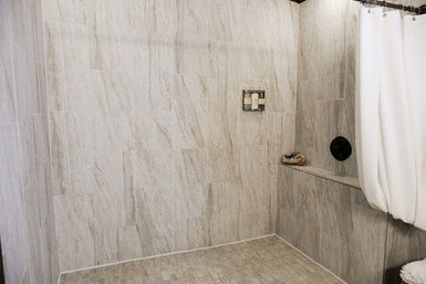 Modern walk-in shower with light gray stone-look wall and floor tiles, built-in bench, white shower curtain, wall-mounted shampoo dispensers and black round shower control