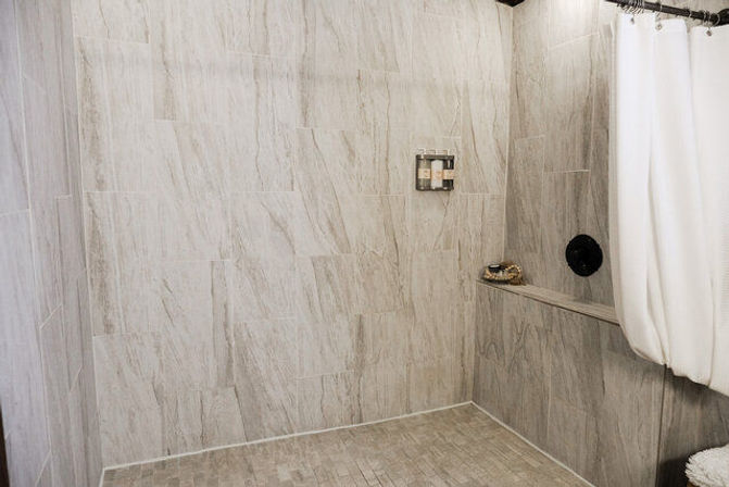 Modern walk-in shower with light gray stone-look wall and floor tiles, built-in bench, white shower curtain, wall-mounted shampoo dispensers and black round shower control