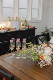 Rustic wooden dining table set for a celebration with six champagne flutes rimmed in pink sugar, golden bubbly, and a lush floral centerpiece, plus a buffet with chilled bottles in an airy dining room.