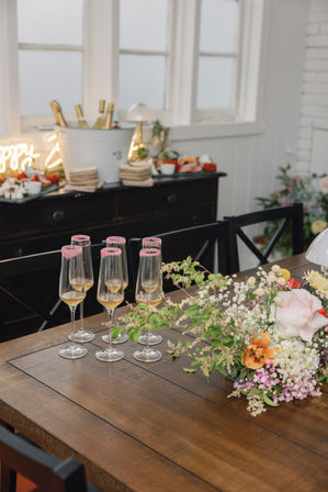 Rustic wooden dining table set for a celebration with six champagne flutes rimmed in pink sugar, golden bubbly, and a lush floral centerpiece, plus a buffet with chilled bottles in an airy dining room.