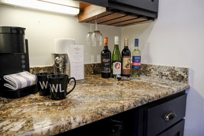 Granite kitchenette countertop with coffee maker, two black mugs, hanging wine glasses and four bottles of wine, ready for coffee or a pour