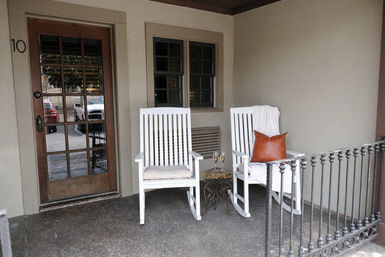 Cozy covered front porch with two white wooden rocking chairs, a small ornate side table holding two wine glasses, a brown leather pillow and draped throw on one chair, glass-paned door marked 10, and black metal railing.