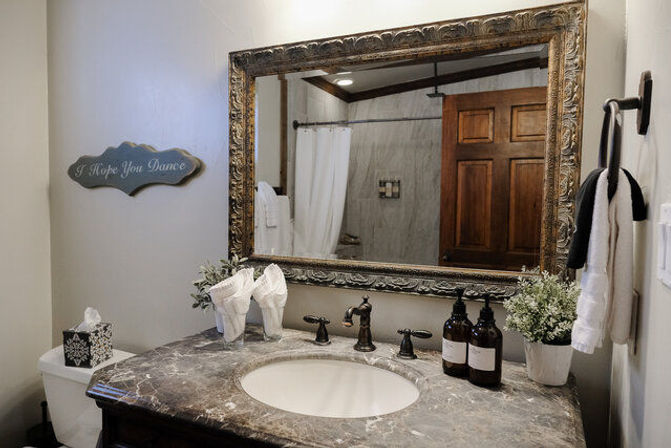 Cozy rustic bathroom vanity with marble countertop and oval sink, ornate gold-framed mirror reflecting wooden door and shower, bronze faucet, amber soap dispensers, folded towel decor and potted plant.