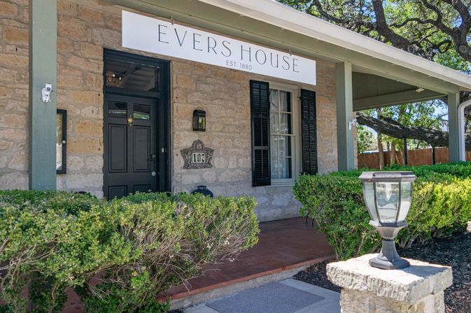 Charming historic limestone house entrance with covered porch, black front door and shuttered window, trimmed hedges and a decorative lantern on a stone post.