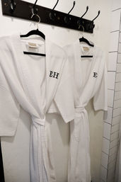 Two monogrammed white waffle-weave spa robes on black hangers, tied at the waist and hanging from wall hooks beside white subway tile in a bathroom.