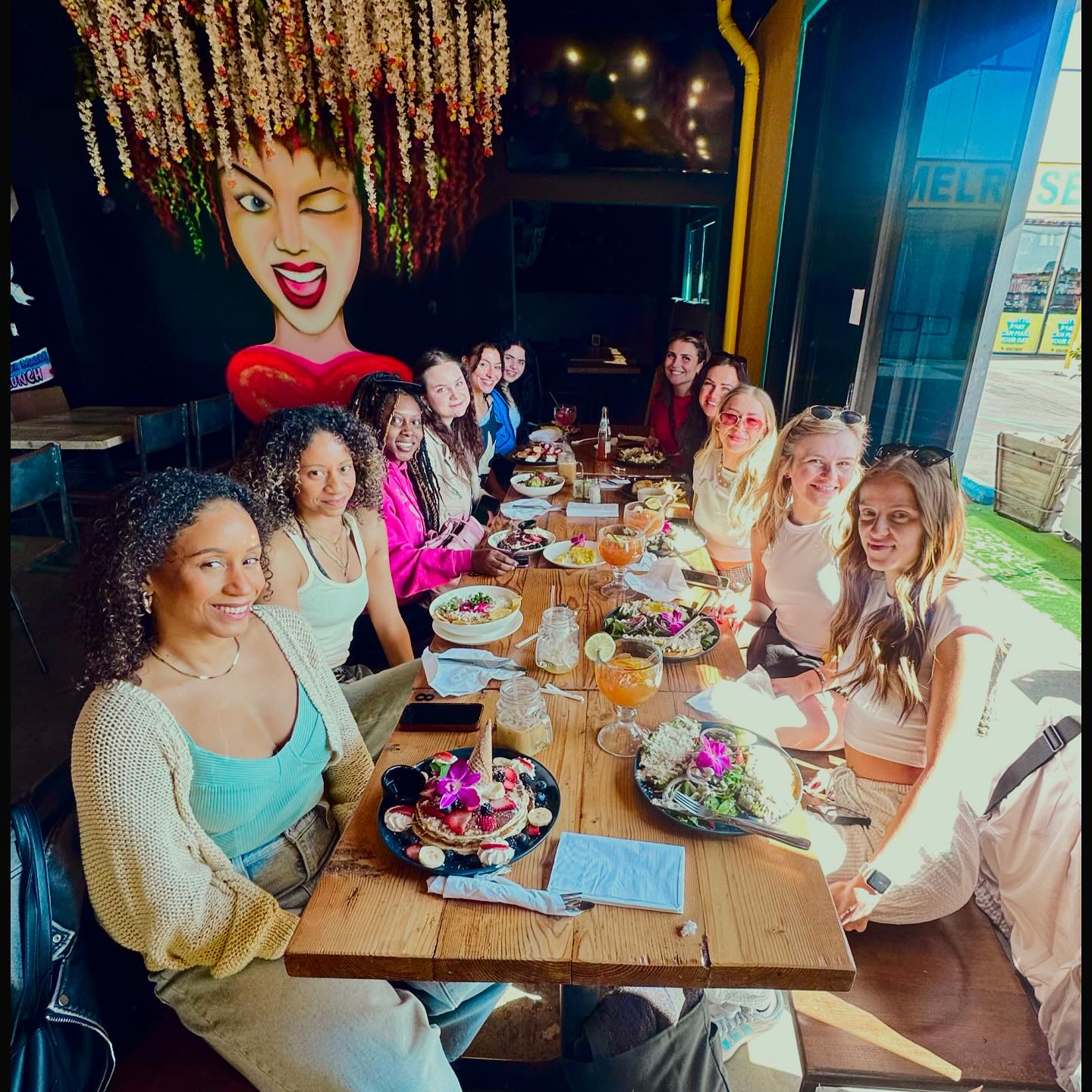 Group of friends enjoying colorful brunch at a sunlit coastal cafe — long wooden table with salads, pancakes topped with edible flowers, cocktails with lime, hanging floral installation and a large winking-face mural, with boardwalk view outside.