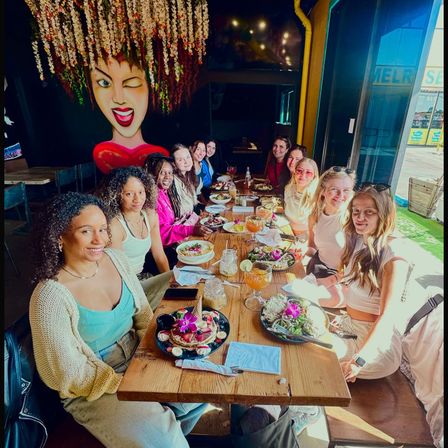 Group of friends enjoying colorful brunch at a sunlit coastal cafe — long wooden table with salads, pancakes topped with edible flowers, cocktails with lime, hanging floral installation and a large winking-face mural, with boardwalk view outside.