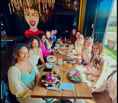 Group of friends enjoying colorful brunch at a sunlit coastal cafe — long wooden table with salads, pancakes topped with edible flowers, cocktails with lime, hanging floral installation and a large winking-face mural, with boardwalk view outside.