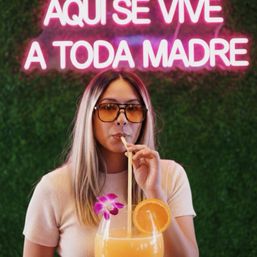 Person wearing sunglasses sipping an oversized orange cocktail garnished with an orchid and orange slice, posed against a green hedge wall under a pink neon sign reading AQUI SE VIVE A TODA MADRE.