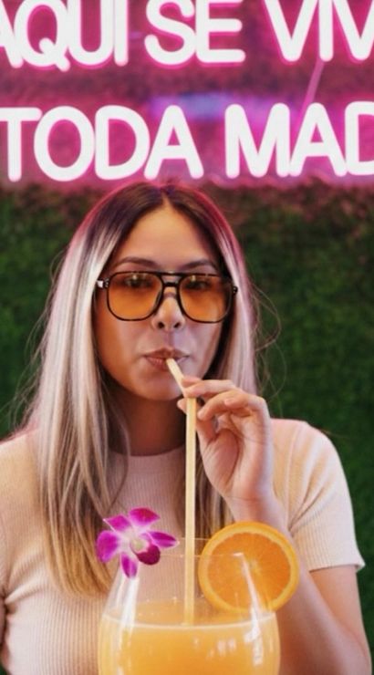 Person wearing sunglasses sipping an oversized orange cocktail garnished with an orchid and orange slice, posed against a green hedge wall under a pink neon sign reading AQUI SE VIVE A TODA MADRE.